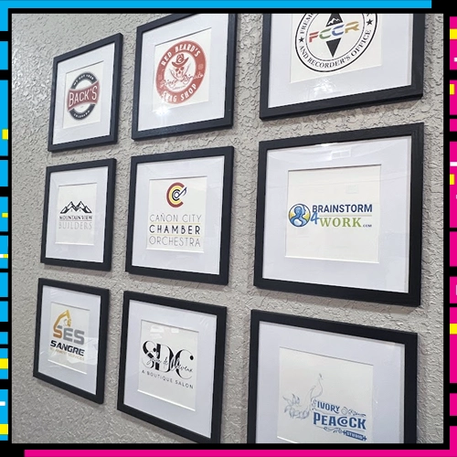 A variety of logos that DARE Print & Sign Co have designed for their clients shown in frames hanging on the office wall.